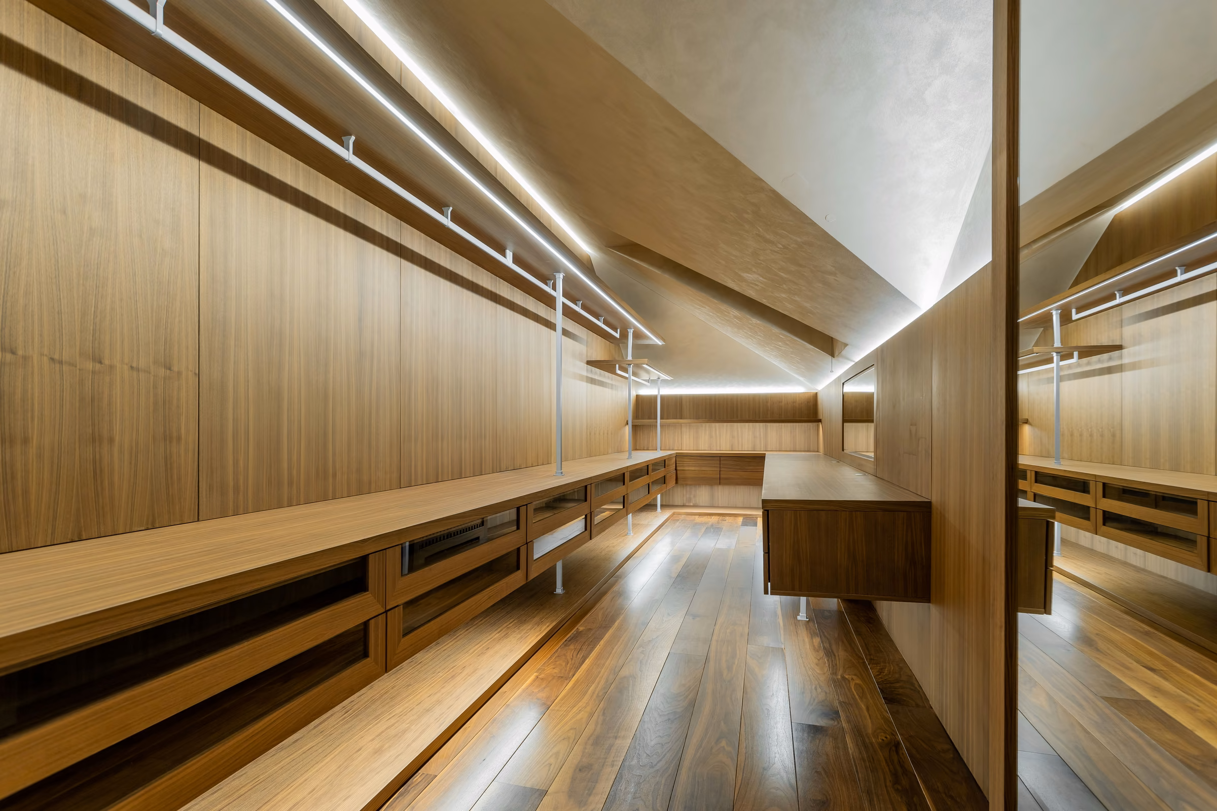 Closet revestido de nogueira com gaveteiros e iluminação linear no forro inclinado